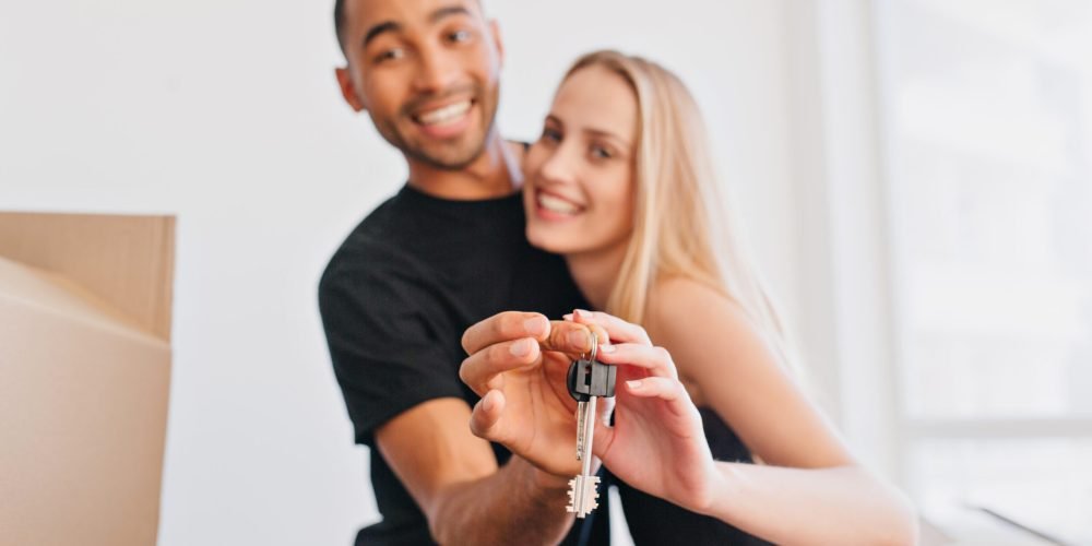 Smiling couple bought new house, holding keys for new home, moving in, packing boxes, relocating. Focus on keys. People, white wall, big window and box on background.